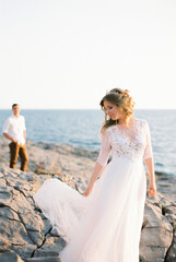 Fototapeta na wymiar Bride lifts the hem of her dress on the rocks by the sea against the background of the groom