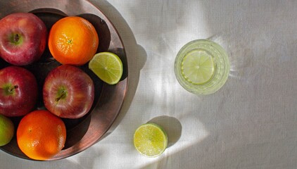 A clay plate with fruit, a glass of water and lemon on a white linen tablecloth in natural light with sunny shadows.