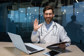 Portrait of successful mature doctor inside modern clinic, man working with laptop looking at camera and smiling waving hand greeting gesture