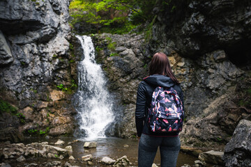 Photo of young and fit brunette woman with backpack looking at the waterfall with strong stream in green forest. Female tourist portrait - hiking to the mountain waterfall in autumn season.