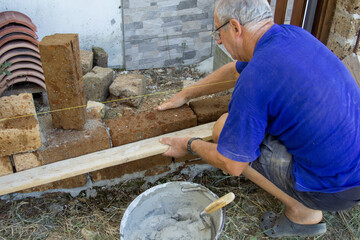 Image of an elderly bricklayer who with a board puts up some tuff bricks. Construction of an...