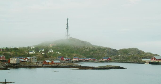 Radio tower on a small hill in the norwegian fjords. A village with classic Norwegian red houses during cloudy, misty morning.
