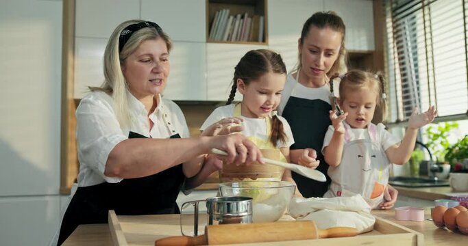 preschooler pretty girl with braids clapping in hands with flour while middle aged granny holding big wooden spoon in hand trying to take flour and sieving on glass bowl. Happy family reunion .