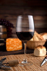 glass of red wine on wooden table with bread and cheese, bottle opener and background wooden barrel with grapes
