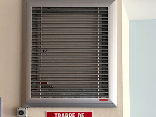 illustration d'une trappe de désenfumage de couleur grise sur un mur avec sa pancarte en dessous de couleur rouge et blanche