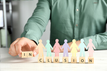 Man making word Inclusion with wooden cubes and paper human figures at table indoors, closeup
