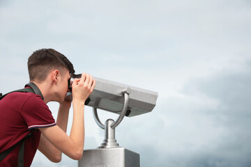Fototapeta na wymiar Teenage boy looking through mounted binoculars under sky. Space for text