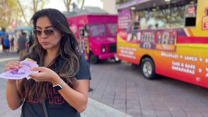 Hispanic woman in the foreground eating food from a food truck - blurred background with several food trucks and people at a festival