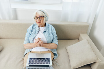 a pleasant mature woman is sitting in a comfortable environment on the couch and listening to a lecture with headphones holding a laptop on her lap learning new things at home