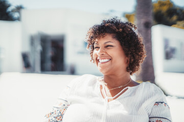 An outdoor portrait of a dazzling happy plus-size Hispanic female with curly hair; a laughing mature caucasian woman with a perfect white smile sitting in a street cafe on a warm sunny day