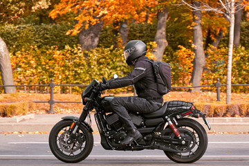 A motorcyclist rides a black retro motorcycle on the road next to the autumn park, the end of the...