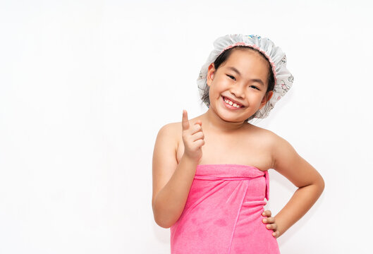 Cute Asian child girl wears shower cap and pink towel, smiling face, one hand on waist and one hand point a finger, isolated cute Asian child girl wears shower cap and pink towel on white background.