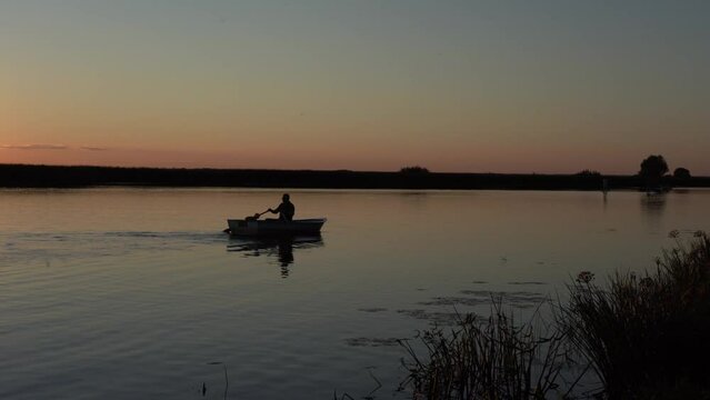 An elderly man with a poodle dog at the stern of the boat rows at dusk in the late evening against the backdrop of sunset