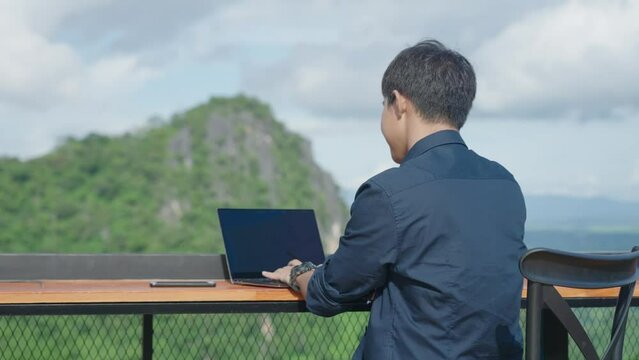 Rear view of asian adult man enjoy workforce life hybrid digital work anywhere, start work, open laptop, log in computer passcode at cozy caf&eacute; workcation place rural green mountain. digital nomad