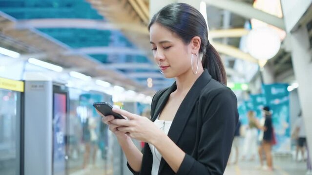 Beautiful asian working woman using smartphone while waiting for the train at Skytrain subway platform, convenient city lifestyle, smart business attire outfit, clients meeting appointment, smart tech