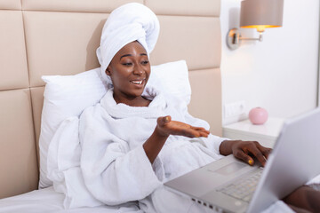 Smiling african woman lying on bed in bathrobe with laptop talking to her friends via video call....