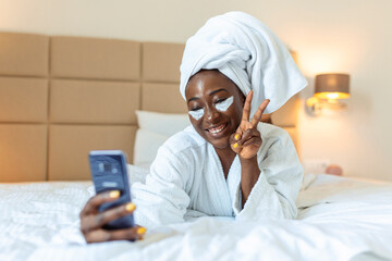 Attractive young black girl in bathrobe and with a towel on her head with eye mask is holding mobile phone,texting and smiling while sitting on a bed. Young African woman on bed