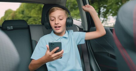Handsome cheerful carefree teen boy in earphones listening music on the backseat during car trip