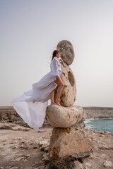 A woman stands on a stone sculpture made of large stones. She is dressed in a white long dress,...