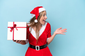 Young blonde woman dressed as Mama claus holding a present isolated on blue background with surprise facial expression