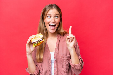 Young caucasian woman holding a burger  isolated on red background intending to realizes the solution while lifting a finger up