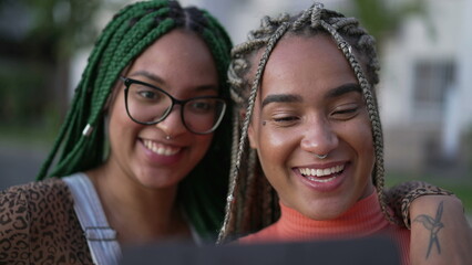 Two happy black adult girls talking to friend on tablet video. Young women with box braids hairstyle holding device outdoors in urban street. Real life authentic smile and laugh 2