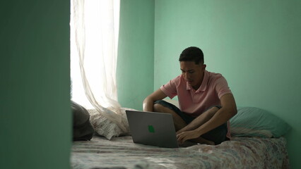 One hispanic young man sitting on bed opening laptop in bedroom bed. Candid South American Brazilian person starting computer searching online
