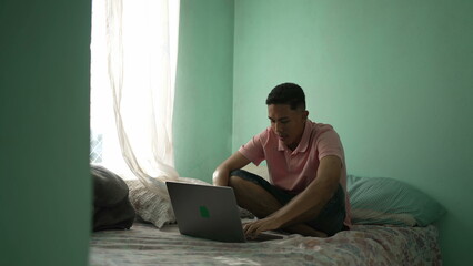 One hispanic young man sitting on bed opening laptop in bedroom bed. Candid South American Brazilian person starting computer searching online