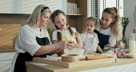 Happy serious concentrated granny teaching granddaughters showing how to beat eggs and adding in glass bowl with flur yo make homemade domestic dough for baking cookies biscuits ay home.