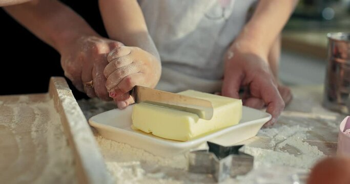 Slow motion selective focus on kid's hands with woman's holding knife in hands trying tu cut butter on white plate. Cooking baking process concept.