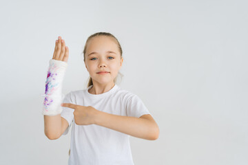 Portrait of cheerful little girl showing with index finger to broken hand wrapped in white plaster bandage with colorful draw looking at camera. Concept of child insurance and healthcare