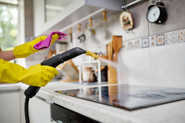 A woman processes the surface of an electric stove with a steam generator while holding a napkin in her hand. Cleaning the kitchen.