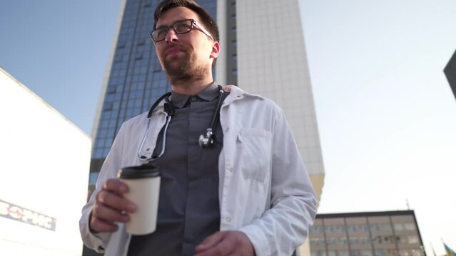 Caucasian male doctor walking and drinking take-away coffee. Male doctor drink hot beverage, walking forward outside hospital building. Physician drinks coffee to go while walking around clinic.