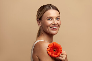 Skin Care Woman Posing with Red Flower. Caucasian Model Holding Flower and Looking At Camera. Female With Nude Makeup, Smooth, Hydrated And Glowing Skin. Natural Beauty On Beige Background  - Powered by Adobe