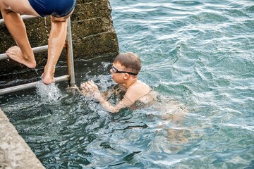Schoolboy climbs up pier on metal ladder against seascape. Waves hit stone pier. Boy enjoys...