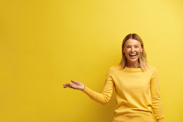Laughing Woman Pointing Palm Away. Blonde Girl Wearing Casual Clothes Gesturing with Smile on Face at Empty Space for Your Advertisement. Indoor Studio Shot Isolated on Yellow Background  - obrazy, fototapety, plakaty