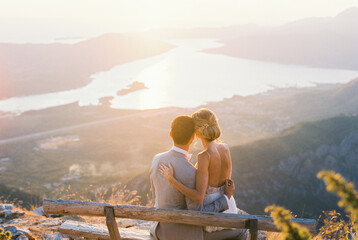 Bride sits on groom lap on a bench on the mountain. Back view