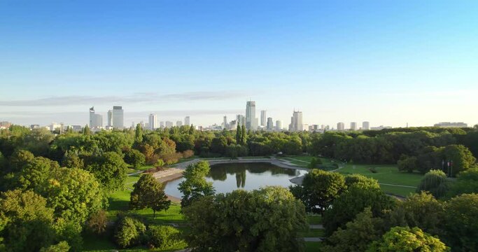 City park with the city center in the background. Lake in the city park with green nature trees during summer morning. Aerial view of offices in skyscrapers over the panorama of Warsaw in a urban park