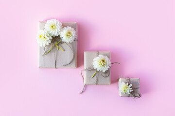 Three gift boxes in gray craft paper decorated with white dahlia flowers on a pink background for the holiday.