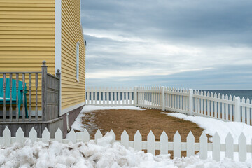 A yellow country style wooden building with a white picket fence attached and enclosing the boundary of a yard overlooking the blue ocean with a cloudy sky. The house has a wood cape cod siding. 