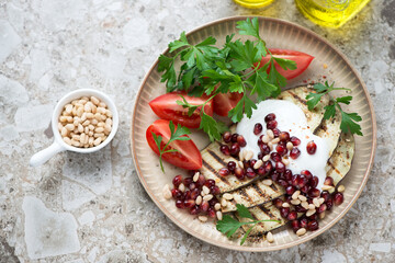 Slices of grilled aubergine with pomegranate, pine nuts, yogurt, tomato wedges and parsley, high angle view on a light-brown granite background, horizontal shot