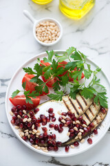 Grilled eggplant with pomegranate seeds, yogurt, tomatoes and fresh parsley, vertical shot on a white marble background, elevated view