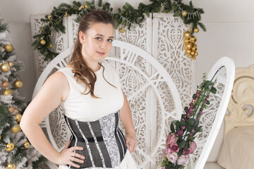 Pretty woman plus-size in a white dress and a black corset against the background of Christmas decorations
