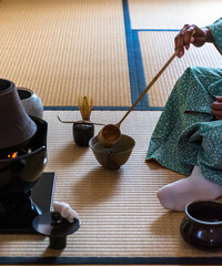 Black woman guest hands using a Hishaku bamboo ladle to pour hot water into chawan tea bowl filled...
