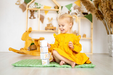 a little baby girl swinging stands a pyramid or a tower of wooden cubes in a playhouse and smiles, a happy child in a yellow dress