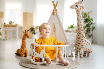 a little child girl in a yellow dress plays with a giraffe at home with a wigwam in a bright room and smiles, happy childhood © Any Grant