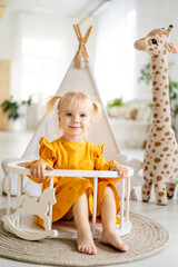 a little child girl in a yellow dress plays with a giraffe at home with a wigwam in a bright room and smiles, happy childhood © Any Grant