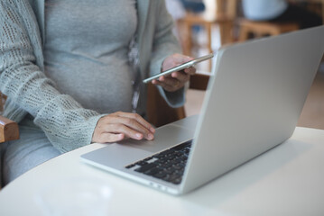 Pregnant business woman sitting at table using mobile phone and working on laptop computer at cafe. pregnant woman online shopping via mobile app, close up