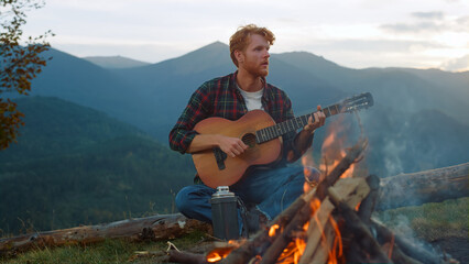 Thoughtful traveler play guitar outdoors. Guy sit by mountains nature campfire.