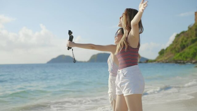 Two Asian female bloggers are taking pictures with action cam to show off to their friends on social media during a beach trip on an island in the sea, concept of freedom, travel, adventure
